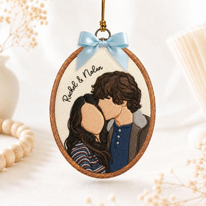 An oval-shaped embroidery hoop ornament featuring a thread-stitched portrait of two people. The text "Rachel & Nolan" is embroidered in black. The hoop is framed in wood and topped with a light blue satin ribbon bow for hanging.
