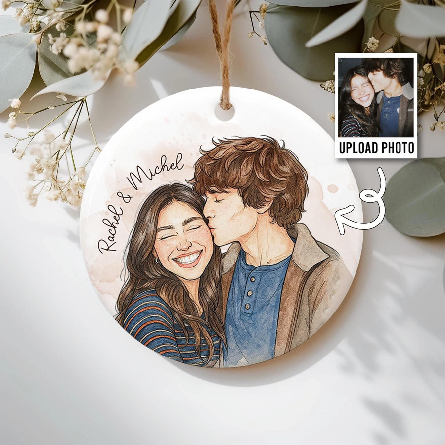 A round, white ceramic ornament with a watercolor illustration of a couple kissing. The ornament has a string for hanging and the names "Rachel & Michel" are written on the side. A small photo is shown with the text "UPLOAD PHOTO".