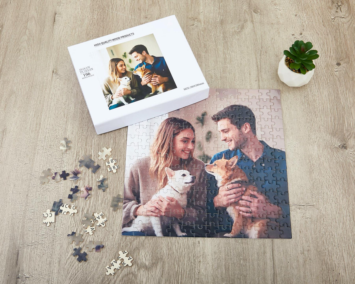 A square jigsaw puzzle featuring a photograph of a person with long hair and a person with short hair, each holding a small dog. The puzzle is partially assembled next to its white box, which displays the same image and text.