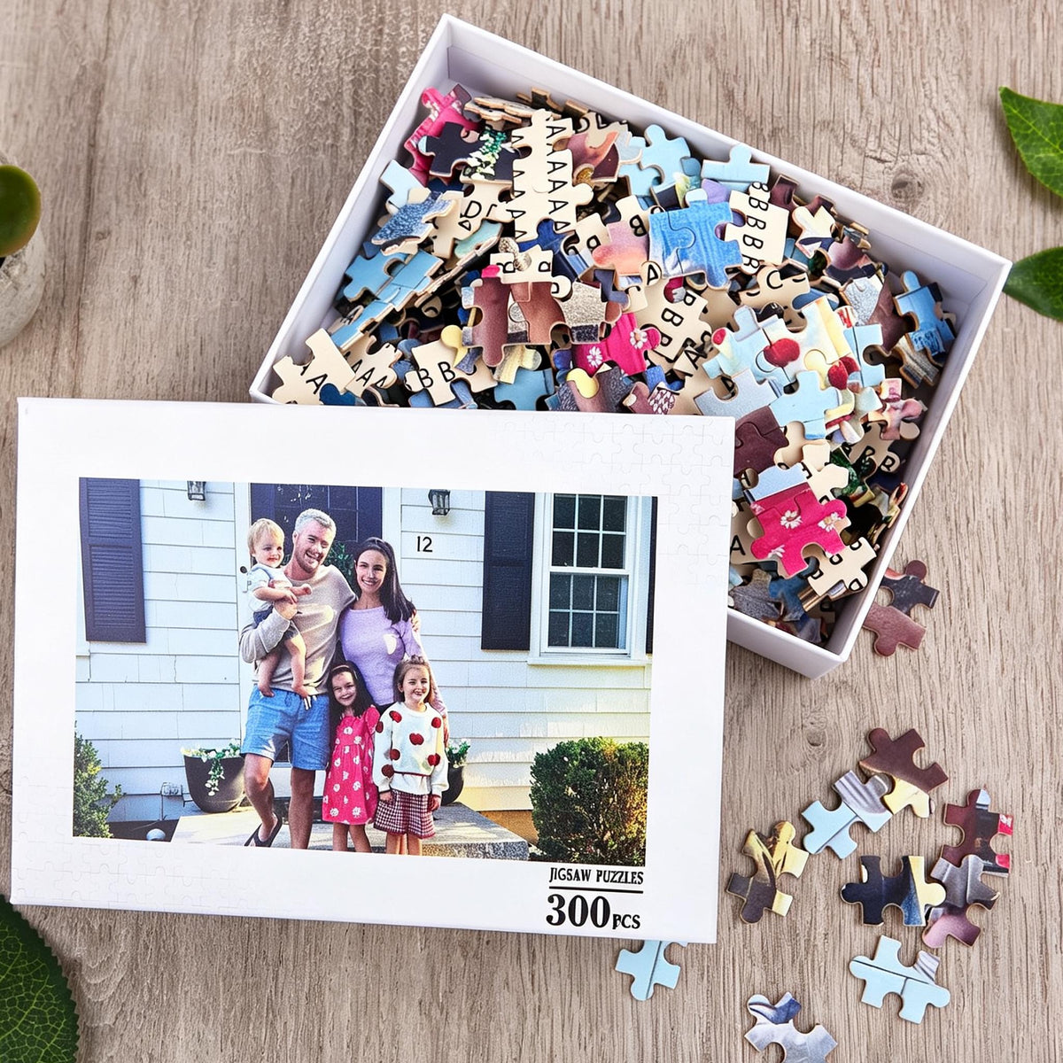 A 300-piece jigsaw puzzle in a white box. The box lid features a photograph of a family of four standing in front of a white house with the number 12 visible. The box is filled with loose, colorful puzzle pieces with letters on the back.