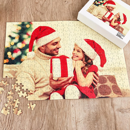 A 300-piece jigsaw puzzle featuring a photograph of an adult and a child wearing red Santa hats, holding a white gift box with a red ribbon. The background shows a blurred Christmas tree with lights. The puzzle box is visible in the corner.