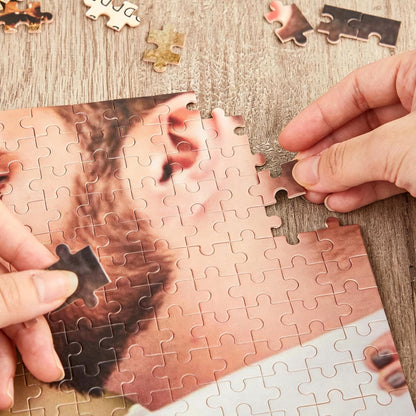A jigsaw puzzle featuring a photographic print of a person's face with dark hair and skin tones. The puzzle is partially assembled with several loose pieces scattered nearby, showing interlocking cardboard edges and a smooth surface finish.