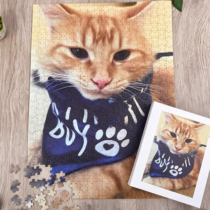 A jigsaw puzzle featuring a ginger cat wearing a dark blue bandana with white paw print graphics and text. The puzzle is partially assembled, with a white box showing the completed image and several loose puzzle pieces nearby.