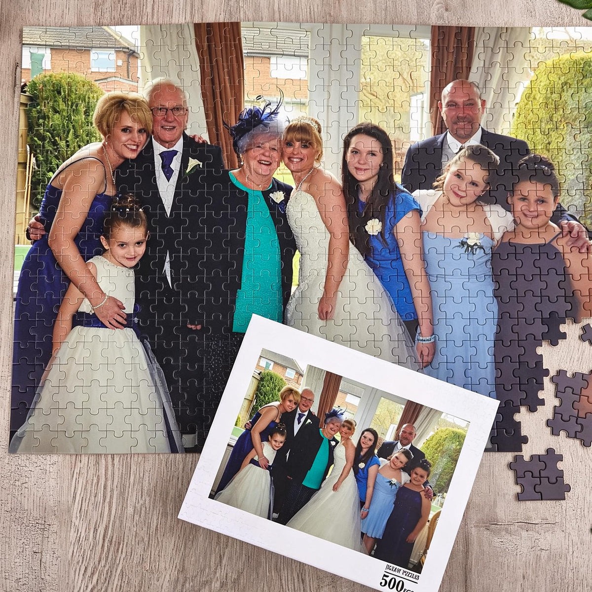 A 500-piece jigsaw puzzle featuring a group portrait of a wedding party. The image shows a bride, groom, and guests in formal attire, including blue and white dresses and suits. A smaller reference print of the same photo is included.