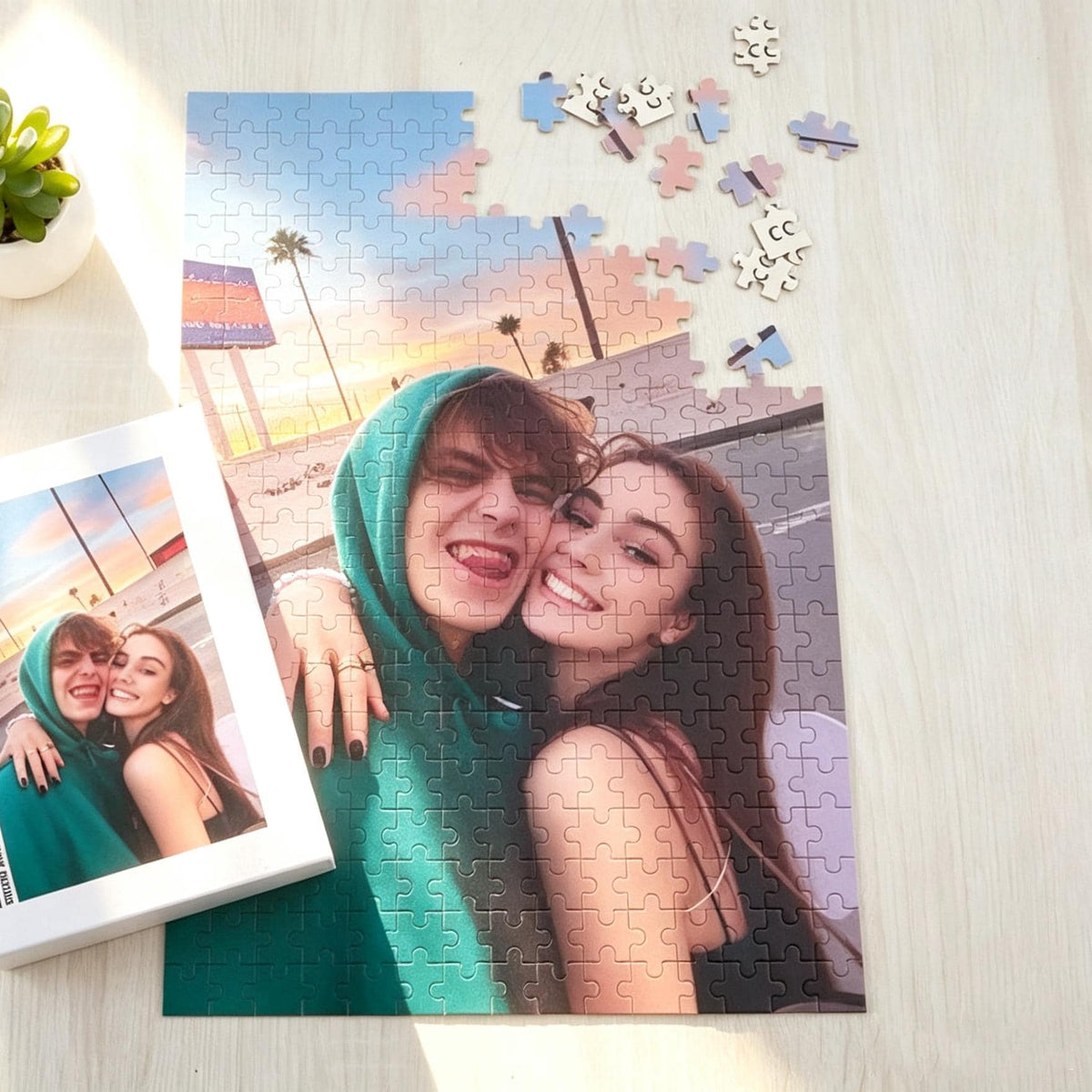 A custom jigsaw puzzle featuring a photograph of two people embracing outdoors at sunset. One person wears a green hooded sweatshirt, while the other has long dark hair. A small box showing the same image is placed next to the puzzle.