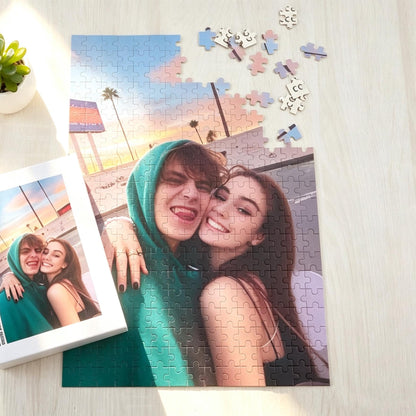A custom jigsaw puzzle featuring a photograph of two people embracing outdoors at sunset. One person wears a green hooded sweatshirt, while the other has long dark hair. A small box showing the same image is placed next to the puzzle.