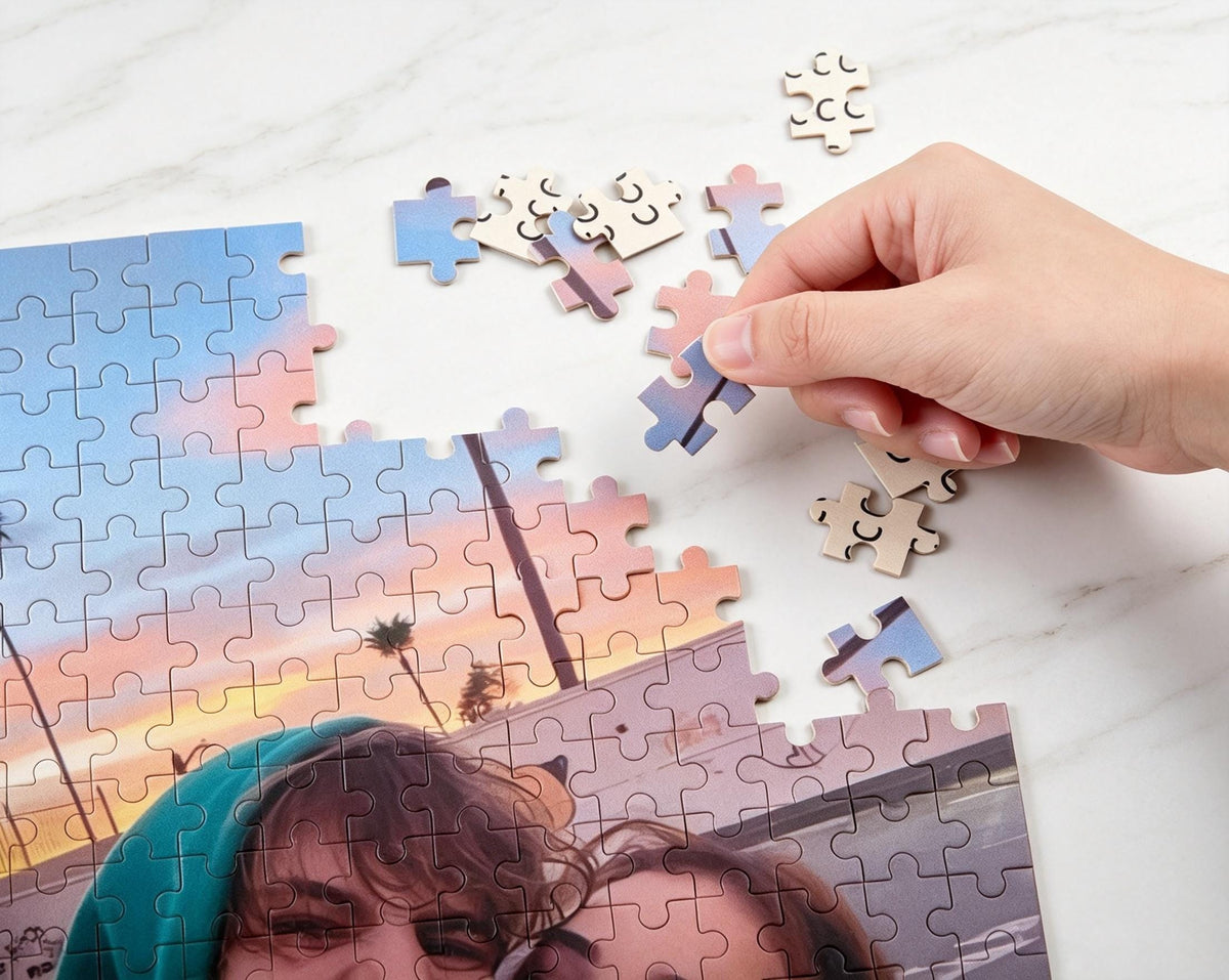 A jigsaw puzzle featuring a sunset beach scene with palm trees and two people in the foreground. The puzzle is partially assembled, with several loose pieces scattered nearby and one piece being placed into the main image.