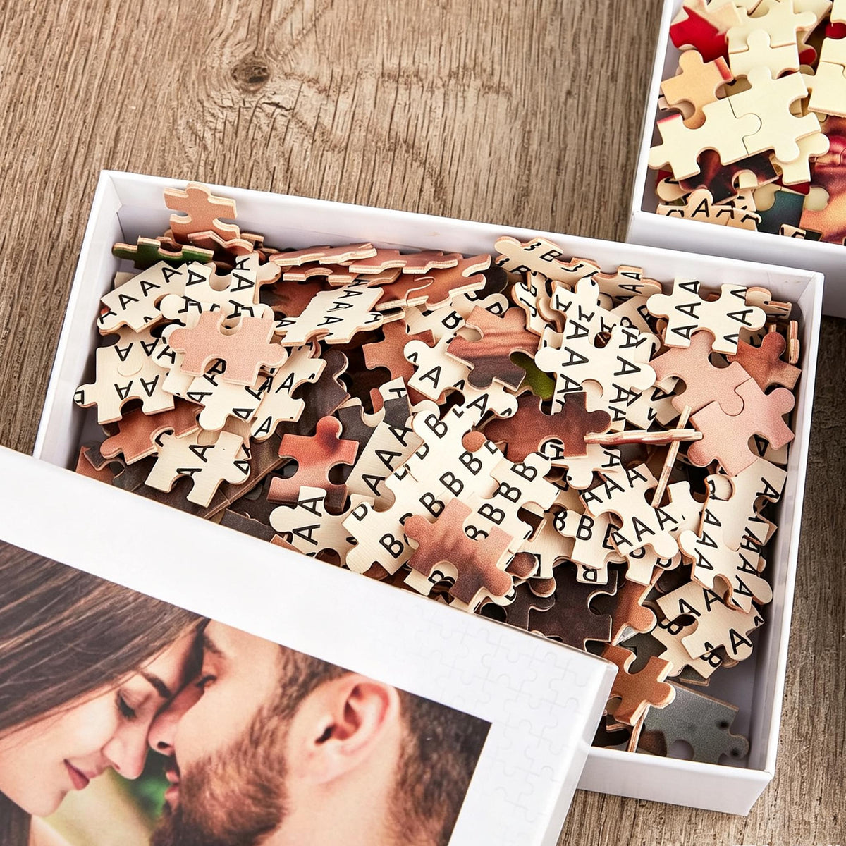 A white cardboard box filled with loose jigsaw puzzle pieces, many featuring the printed letters A and B on the back. The box lid displays a photographic portrait of a couple in close proximity, with a second box visible nearby.