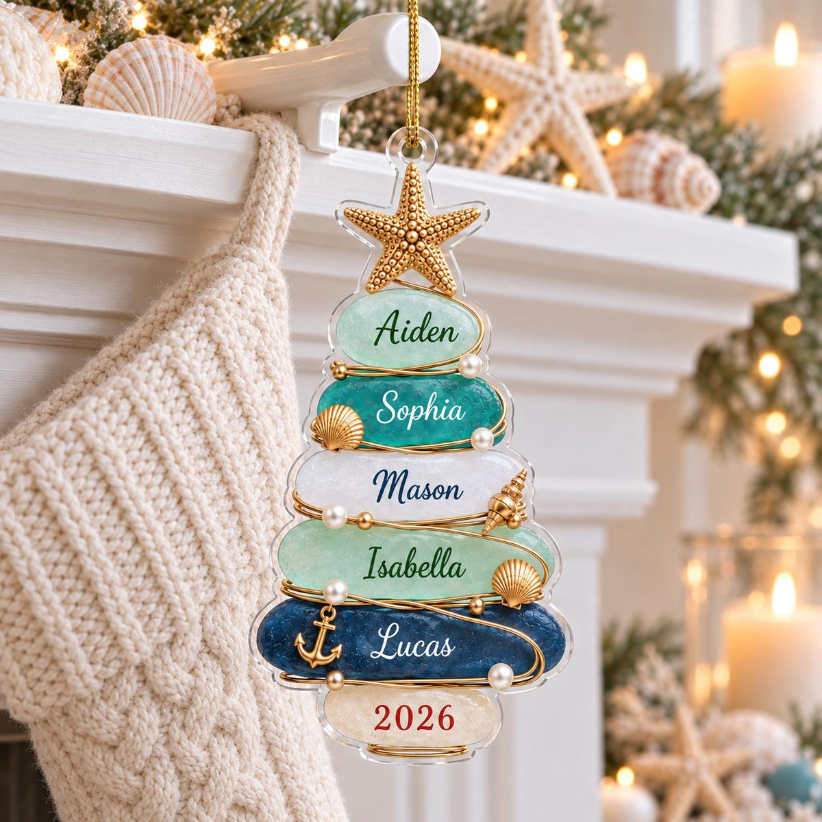 A Christmas tree-shaped ornament made of stacked, smooth, multi-colored stones in shades of blue, green, and white. The ornament features a gold starfish topper, gold wire accents, small pearls, seashells, and an anchor charm. Names Aiden, Sophia, Mason, Isabella, Lucas, and the year 2026 are printed on the stones.