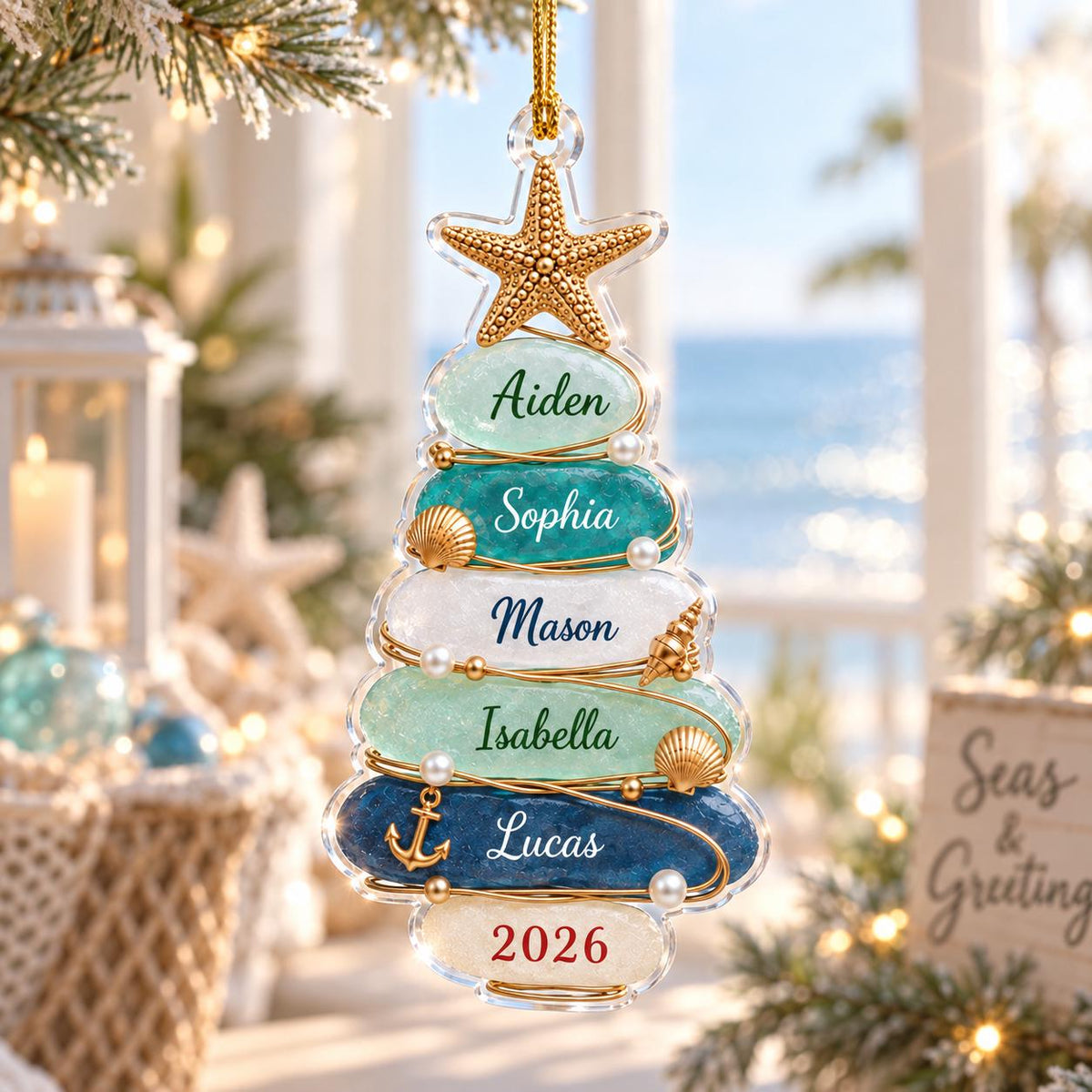 A clear, tree-shaped Christmas ornament featuring stacked, textured glass-like layers in shades of blue, teal, and white. The ornament is topped with a gold starfish and decorated with small pearls, shells, and an anchor, with names and 2026 printed.