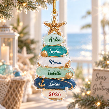 A clear, tree-shaped Christmas ornament featuring stacked, textured glass-like layers in shades of blue, teal, and white. The ornament is topped with a gold starfish and decorated with small pearls, shells, and an anchor, with names and 2026 printed.