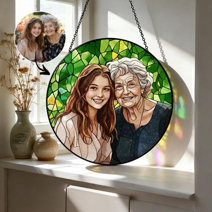 A round stained glass art piece featuring a portrait of two smiling women. The background is a mosaic of green glass. The artwork is suspended by a black chain. A smaller photo of the women is in the upper left corner.