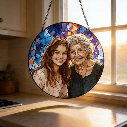 A round stained glass window featuring a portrait of two smiling women. The background is a mosaic of blue, purple, and orange glass. The window is framed in black and suspended by a chain. The women are depicted with realistic features and warm expressions.
