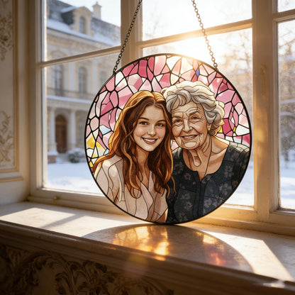 A round stained glass art piece featuring two smiling women, set against a pink and white background. The artwork is framed in black and hangs in front of a window with a snowy outdoor scene.