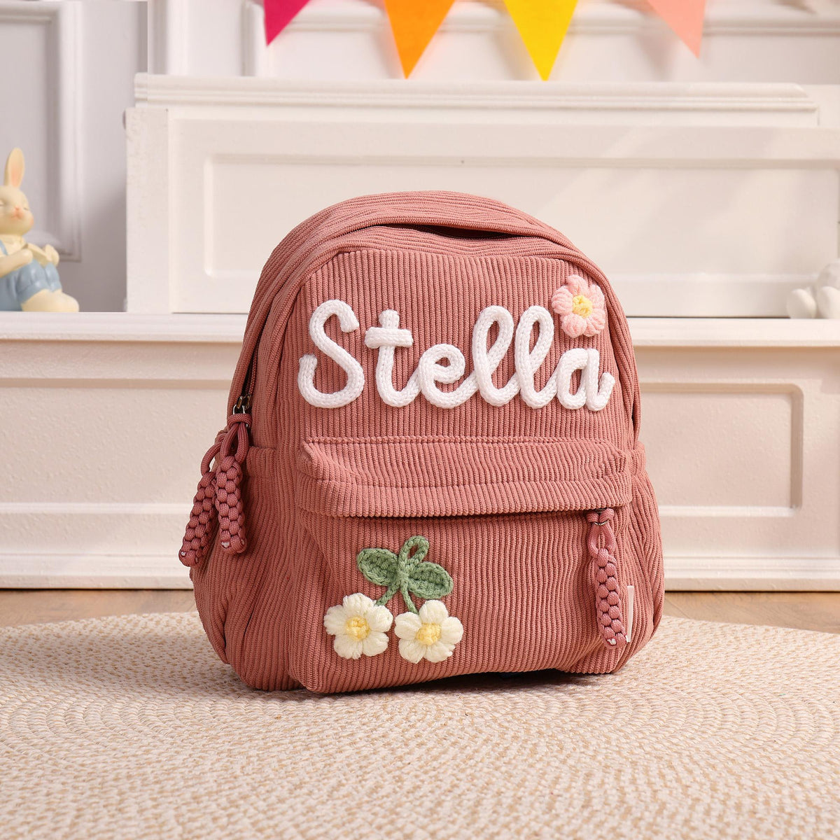 A dusty rose corduroy backpack featuring the name Stella in white embroidered script lettering. The bag includes a small front pocket, a single pink flower appliqu&eacute;, and a green stem with two white flower appliqu&eacute;s at the bottom.