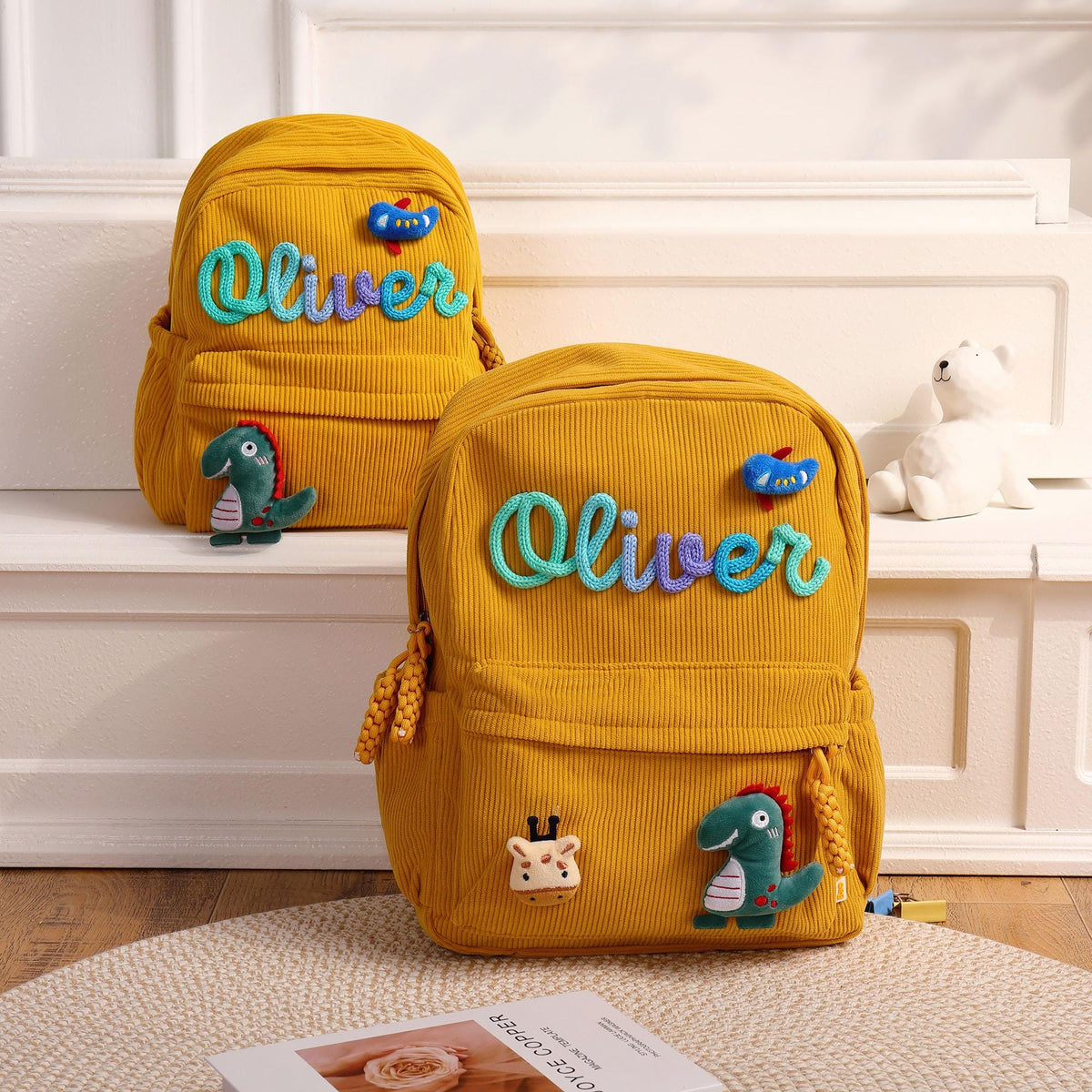 Two mustard yellow corduroy backpacks featuring the name Oliver in a blue and purple script font. Each bag is decorated with a small blue bird patch and a green dinosaur patch. The front pocket includes a giraffe head patch.