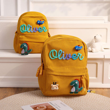 Two mustard yellow corduroy backpacks featuring the name Oliver in a blue and purple script font. Each bag is decorated with a small blue bird patch and a green dinosaur patch. The front pocket includes a giraffe head patch.