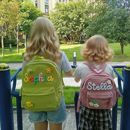 A lime green corduroy backpack with the name Sophia in colorful letters, a bee, a flower, and a duck patch, alongside a dusty pink corduroy backpack with the name Stella in white letters and flower patches on the front pocket.