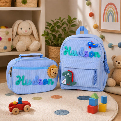 A light blue corduroy backpack and matching lunch bag set, both personalized with the name Hudson in colorful textured lettering. The backpack features a small blue airplane and a green dinosaur patch, while the lunch bag has a lion patch.