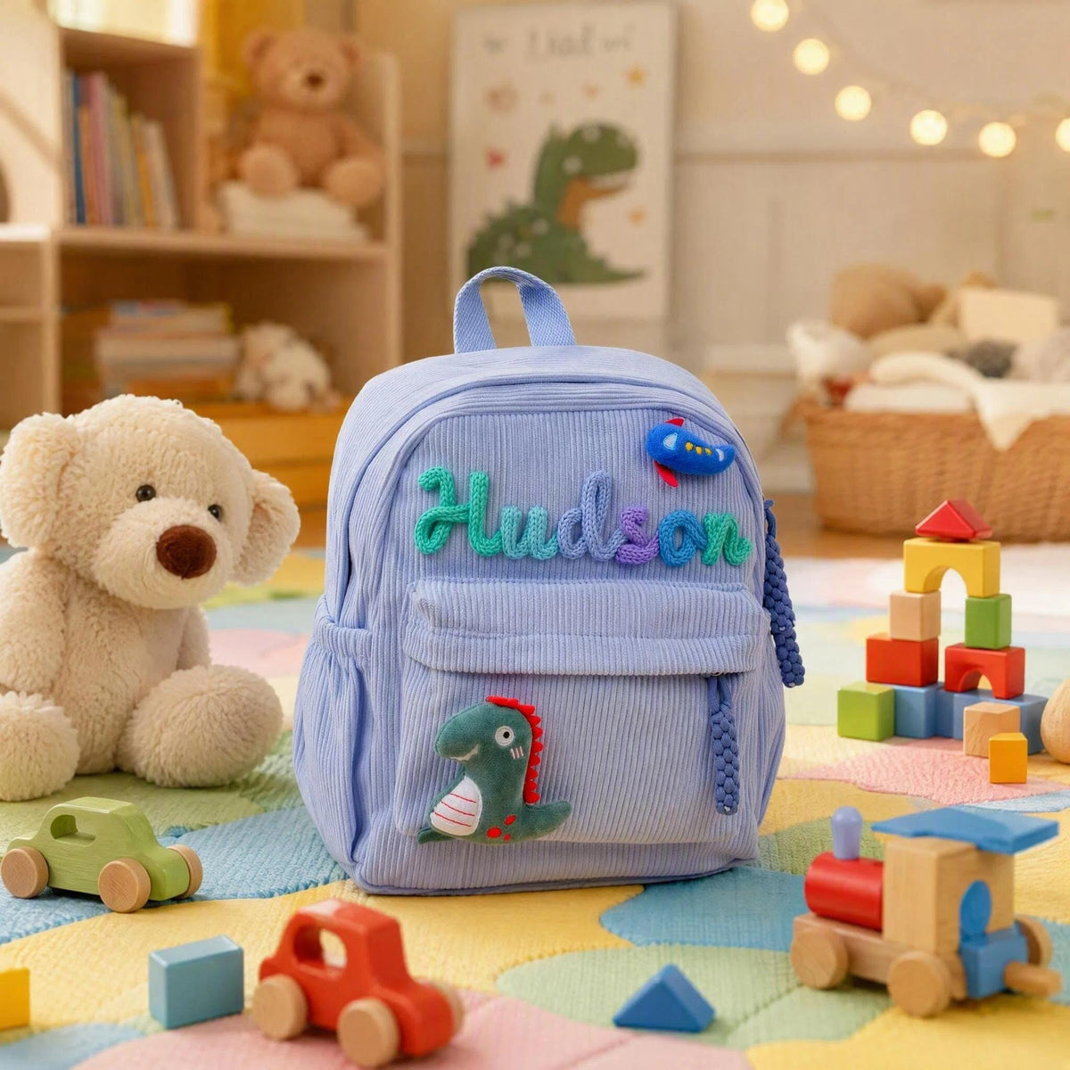 A light blue corduroy backpack featuring the name Hudson embroidered in multi-colored yarn letters across the front pocket. The bag is decorated with a small plush green dinosaur appliqu&eacute; and a blue airplane patch above the name.