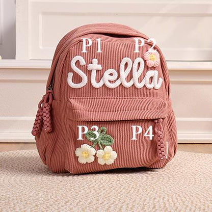 A dusty pink corduroy backpack featuring the white embroidered name Stella, a small pink crocheted flower accent, and a larger green and white crocheted floral applique on the front pocket with braided zipper pulls.
