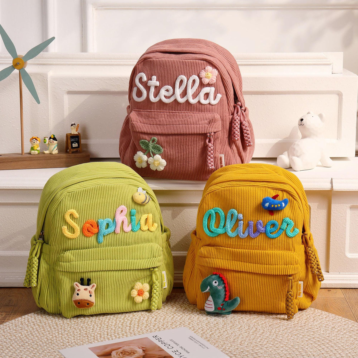 Three corduroy backpacks in dusty pink, lime green, and mustard yellow. Each bag features a front pocket, braided zipper pulls, and embroidered names: Stella with a flower, Sophia with a bee and giraffe, and Oliver with a plane and dinosaur.