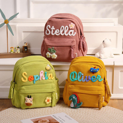 Three corduroy backpacks in dusty pink, lime green, and mustard yellow. Each bag features a front pocket, braided zipper pulls, and embroidered names: Stella with a flower, Sophia with a bee and giraffe, and Oliver with a plane and dinosaur.