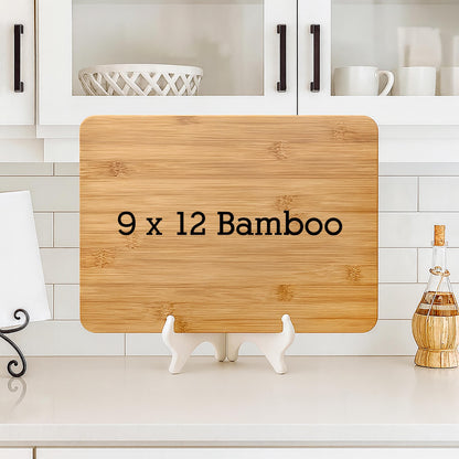 A rectangular bamboo cutting board, measuring 9 x 12 inches, with the text "9 x 12 Bamboo" printed in black. The board is displayed on a white stand against a white tiled background.