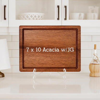A rectangular acacia wood cutting board with a juice groove. The board is 7 x 10 inches and features the text "7 x 10 Acacia w/JG" in white. It is displayed on a clear stand against a white kitchen backdrop.