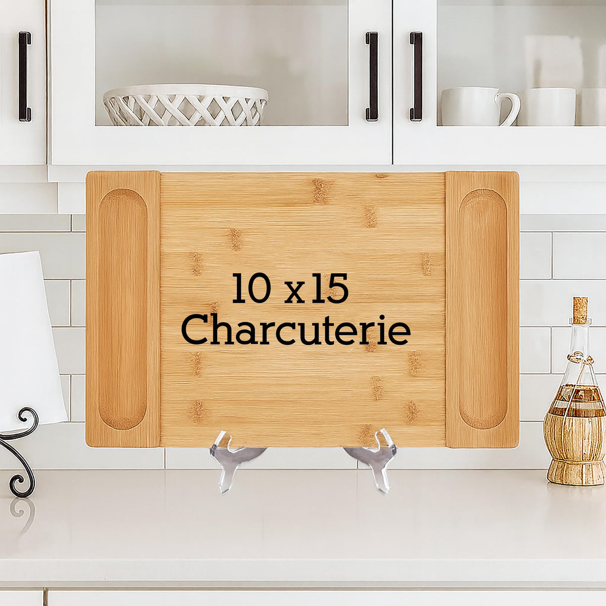 A rectangular bamboo charcuterie board, 10 x 15 inches, with recessed handles. The text "10 x 15 Charcuterie" is printed in black. The board is displayed on a white countertop with a white tile backsplash.