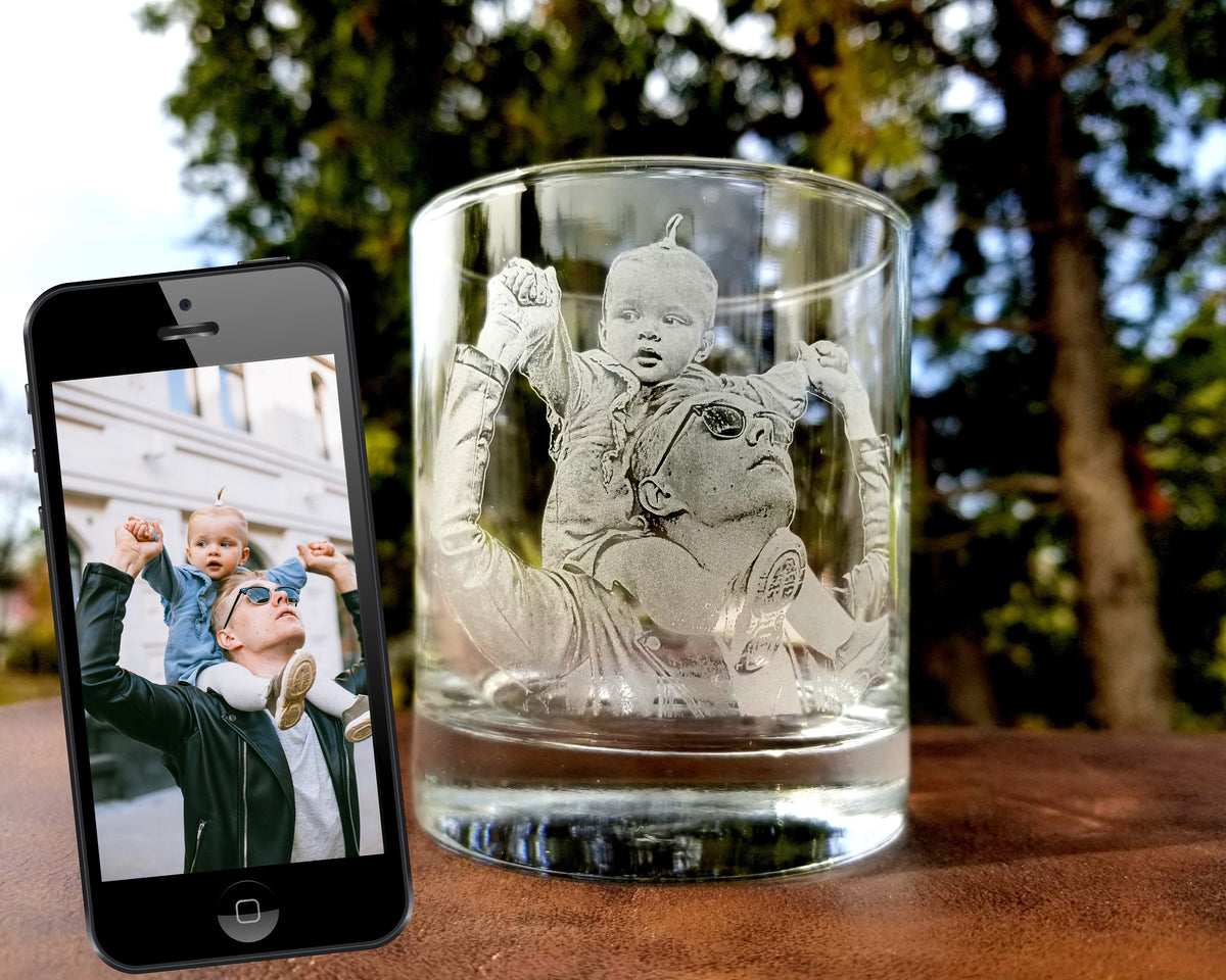 A clear glass with a design etched into the side. The design shows a father and son, with the son sitting on the father's shoulders. The father is wearing sunglasses and the son is looking up.