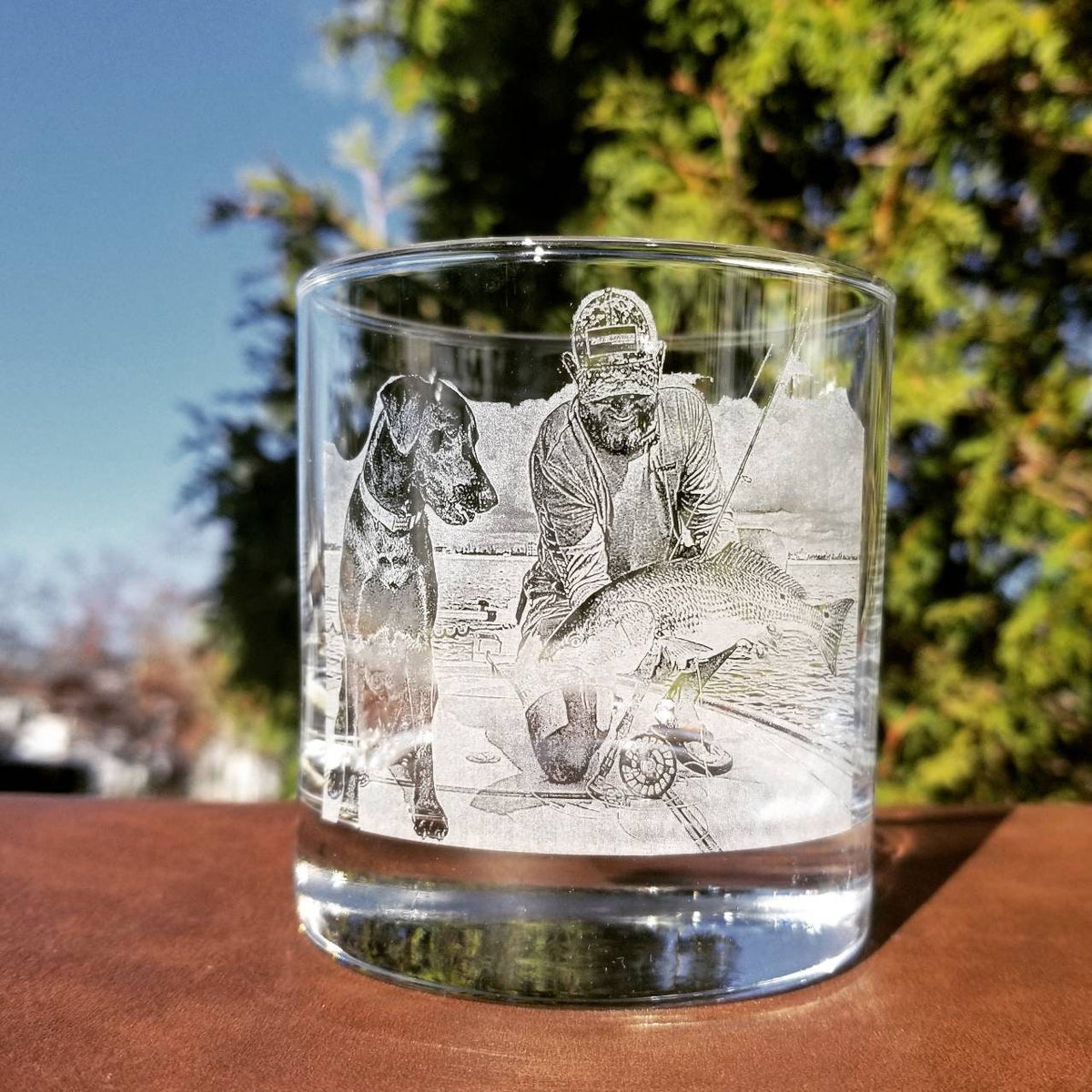 A clear glass tumbler with an etched image of a person holding a fish and a dog standing beside them. The image is in black and white.
