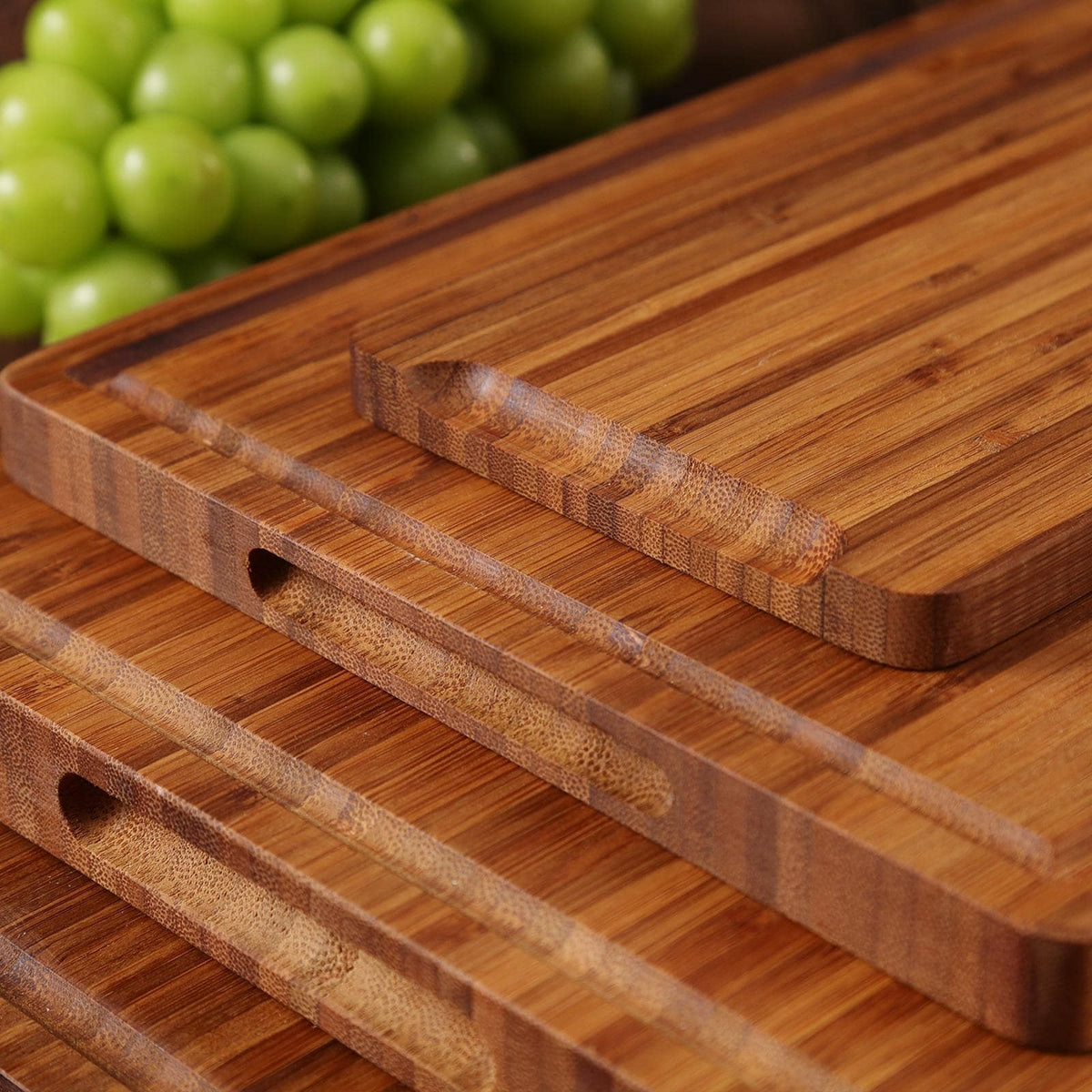 Four wooden cutting boards stacked on top of each other. The boards are made of light brown bamboo and have a smooth, polished surface. Each board has a groove around the edge and a small hole in the center.