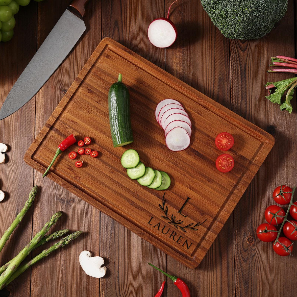 A personalized bamboo cutting board with the name "Lauren" engraved on it. The board is surrounded by various vegetables, including cucumbers, radishes, and tomatoes.