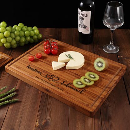 A personalized bamboo cutting board with the names "Kristene" and "Edward" engraved on it, along with the date "EST. DECEMBER 2021". The board is decorated with a heart design and features a cheese wedge, kiwi slices, and cherry tomatoes.