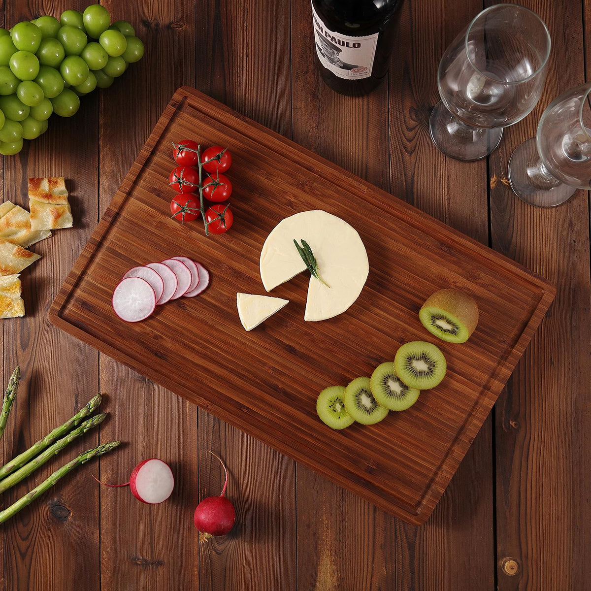 A rectangular brown bamboo cutting board with a variety of food items arranged on it, including sliced cheese, red radishes, cherry tomatoes, kiwi slices, and a sprig of rosemary.