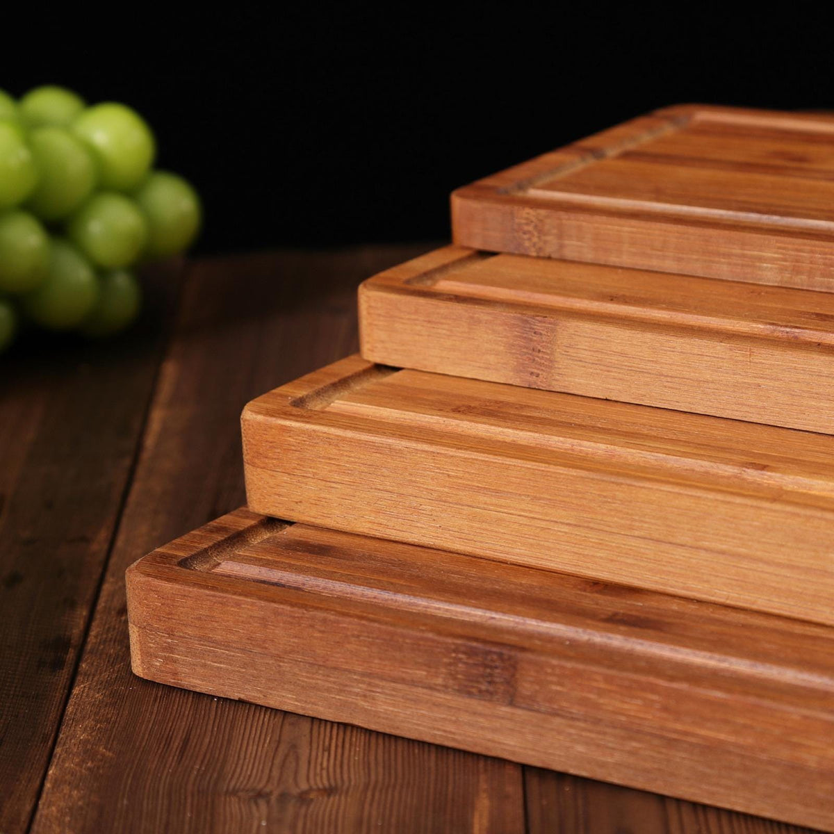 Four wooden cutting boards stacked on top of each other. The boards are made of light brown bamboo and have a rectangular shape. The boards have grooves on the surface to help drain liquids.