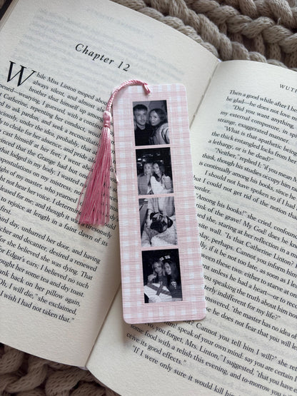 A rectangular bookmark with a light pink and white gingham pattern featuring a vertical strip of four black and white photographs. A light pink tassel is attached to the top center of the bookmark, which is resting on an open book.