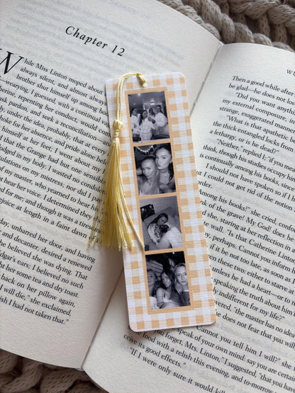A rectangular bookmark with a light orange and white gingham pattern featuring four vertical black and white photographs of people. A pale yellow tassel is attached to the top center hole of the bookmark, which rests on an open book.