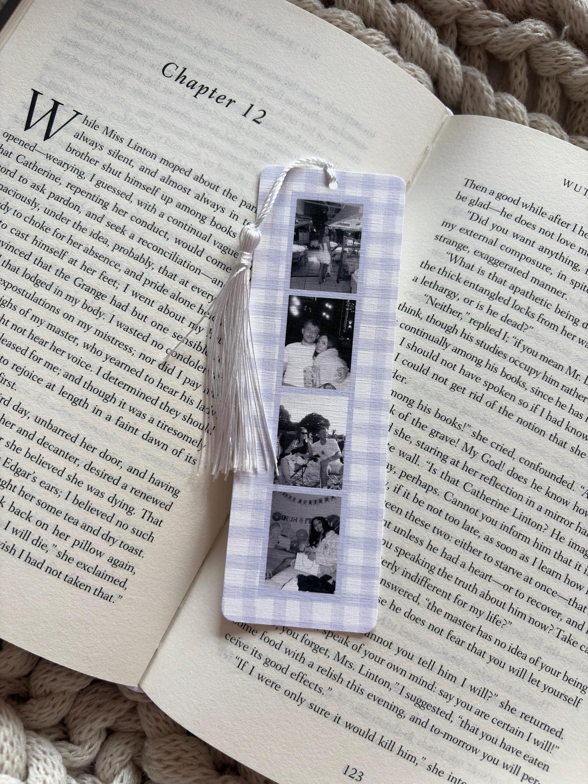 A rectangular bookmark with a light purple and white gingham pattern featuring four vertical black and white photographs of people. A white string tassel is attached to the top of the bookmark, which is resting on an open book page.