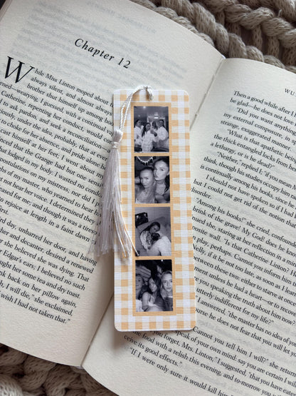 A rectangular bookmark with a light yellow and white gingham pattern featuring a vertical strip of four black and white photographs of people. A white tassel is attached to the top of the bookmark, which is resting on an open book.