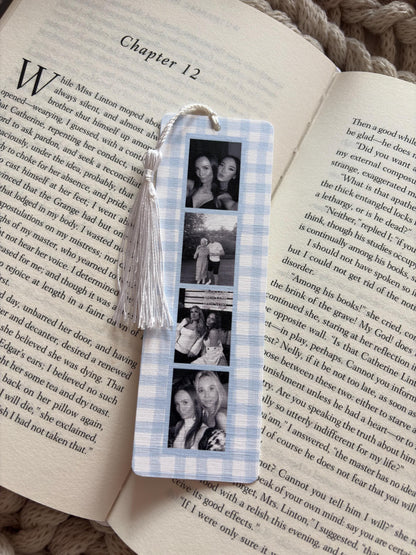 A rectangular bookmark with a light blue and white gingham pattern featuring four vertical black and white photographs of people. A white tassel is attached to the top center hole of the bookmark, which is resting on an open book page.