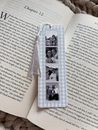 A rectangular bookmark with a light blue and white gingham border, featuring a vertical strip of four black and white photographs. A white cord tassel is attached to the top center of the bookmark, which rests on an open book page.