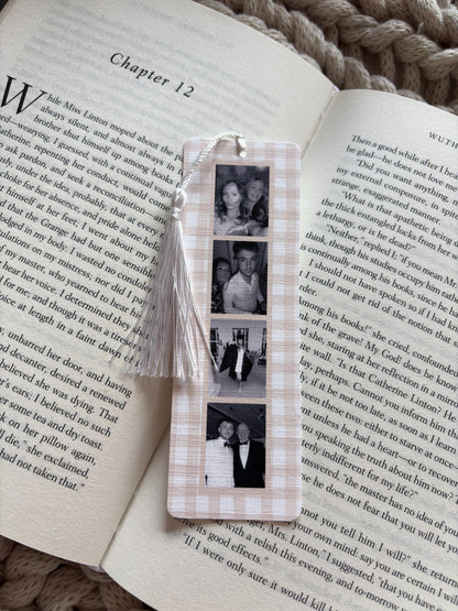 A rectangular bookmark with a light beige and white gingham pattern featuring a vertical strip of four black and white photographs. A white string tassel is attached to the top hole of the bookmark, which is resting on an open book.