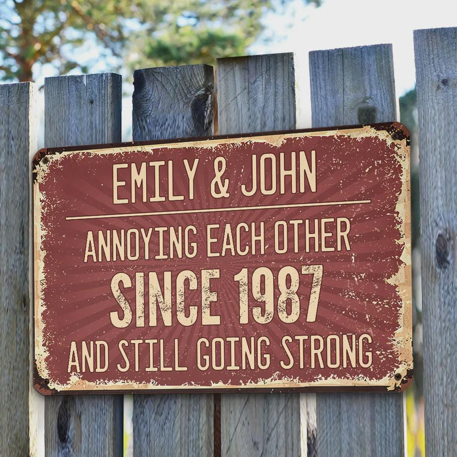 A rectangular, vintage-style metal sign with the text "EMILY & JOHN ANNOYING EACH OTHER SINCE 1987 AND STILL GOING STRONG" in white lettering on a maroon background. The sign is mounted on a weathered wooden fence.