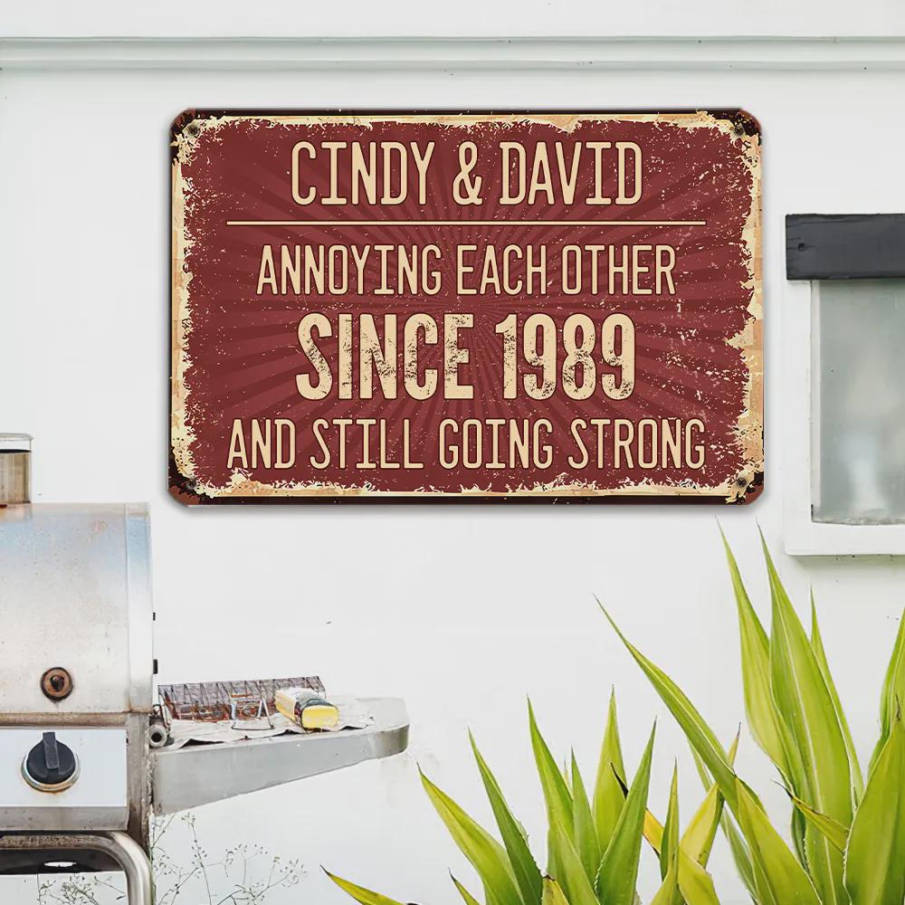 A rectangular, distressed metal sign with a burgundy background and cream-colored text. The sign reads "CINDY & DAVID ANNOYING EACH OTHER SINCE 1989 AND STILL GOING STRONG."
