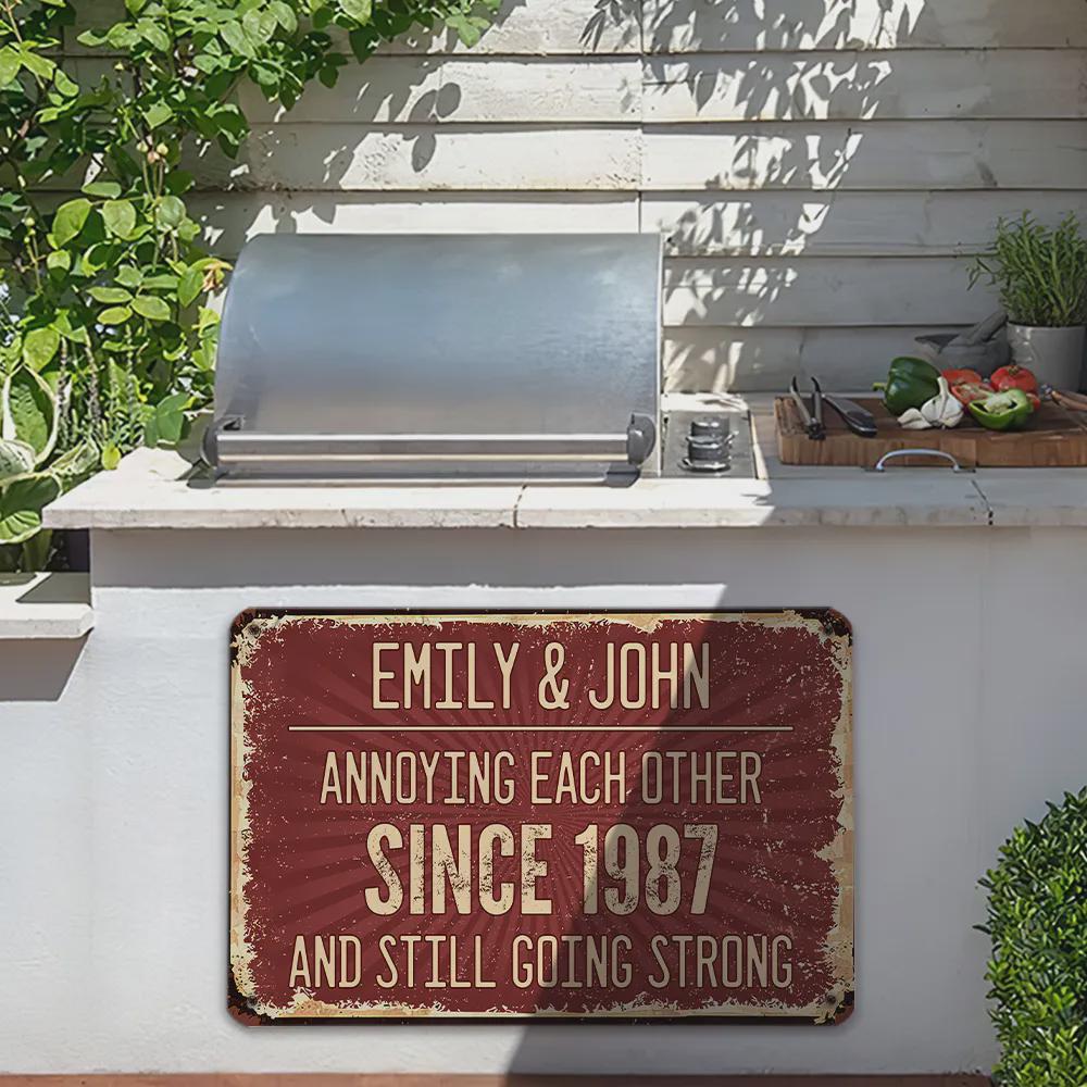 A distressed red metal sign with the text "EMILY & JOHN ANNOYING EACH OTHER SINCE 1987 AND STILL GOING STRONG." The sign is in front of a stainless steel grill and a white concrete structure.