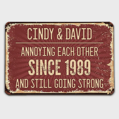 A rectangular, distressed metal sign with a burgundy background and cream-colored text. The sign reads "CINDY & DAVID ANNOYING EACH OTHER SINCE 1989 AND STILL GOING STRONG."