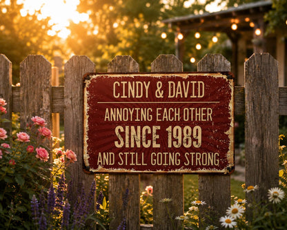 A rectangular, distressed red metal sign with the text "CINDY & DAVID ANNOYING EACH OTHER SINCE 1989 AND STILL GOING STRONG" is mounted on a wooden fence. The background features flowers and a sunset.