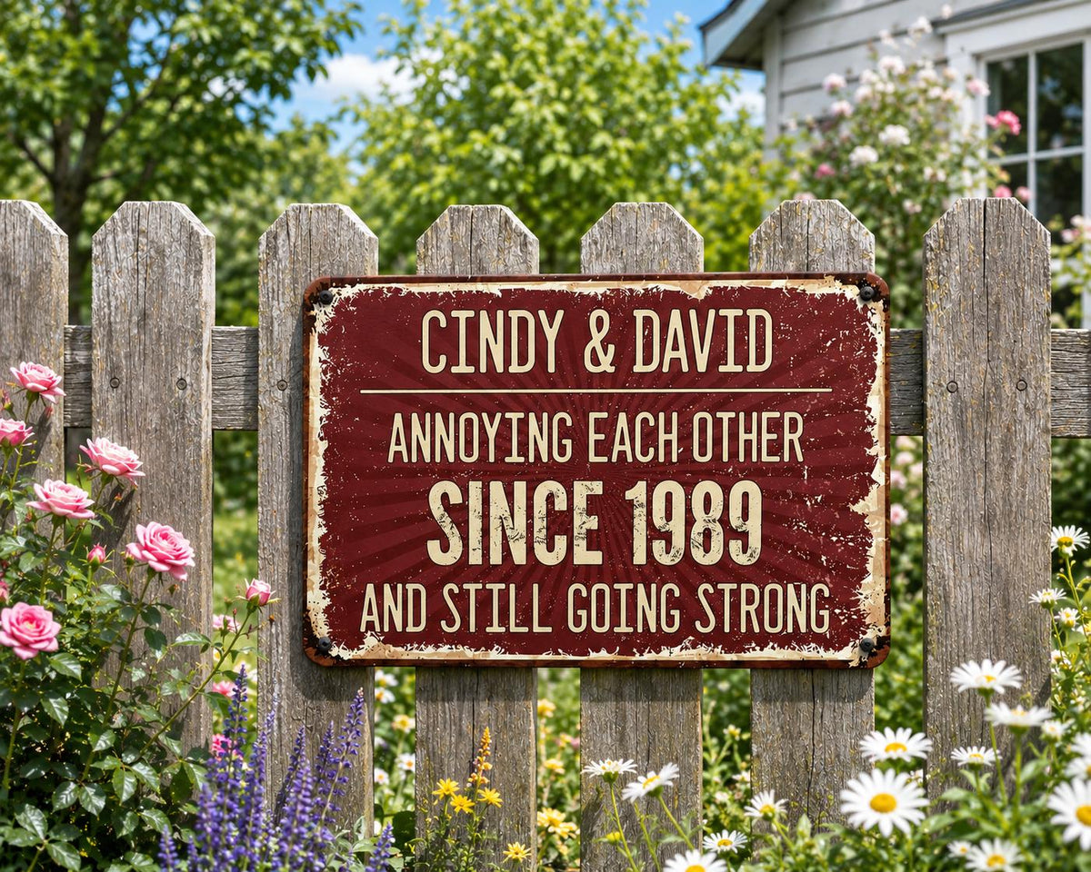A rectangular, distressed burgundy metal sign with white text. The sign reads "Cindy & David Annoying Each Other Since 1989 And Still Going Strong." The sign is mounted on a weathered wooden fence, surrounded by flowers.